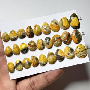 May include: A selection of polished, oval gemstones in yellow, orange, black, and grey. The stones are displayed on a white surface, highlighting their unique patterns and textures. These gemstones are likely for jewellery making or collecting.