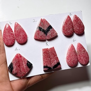 May include: A collection of six pairs of polished gemstones, each with a unique shape and pattern. The stones are primarily a vibrant pink with speckled textures, some featuring black veining. The gemstones are displayed on a white surface.