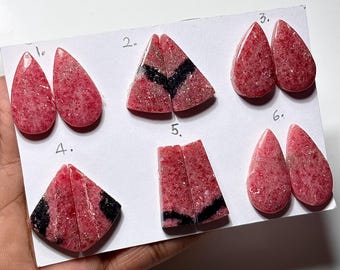 Puede incluir: Una colección de seis pares de piedras preciosas pulidas, cada una con una forma y un patrón únicos. Las piedras son principalmente de color rosa con texturas moteadas, algunas con vetas negras. Las piedras preciosas se muestran sobre una superficie blanca.