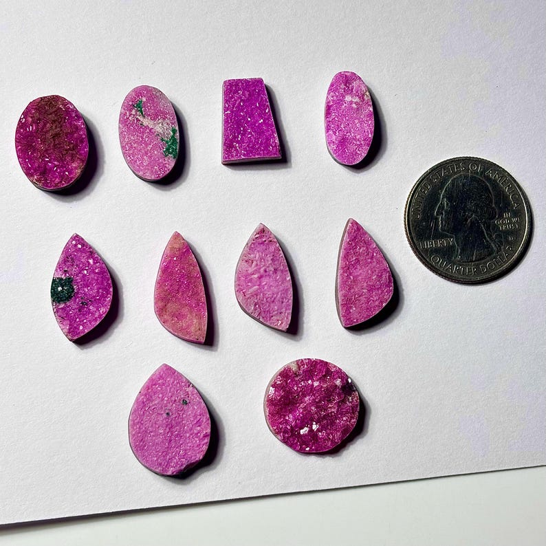 May include: A collection of polished, vibrant pink gemstones in various shapes, including ovals, teardrops, and rectangles. A US quarter is included for size reference. The gemstones have a textured surface and some have green inclusions.