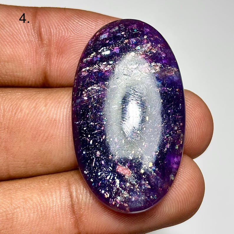 May include: An oval-shaped, polished gemstone with a deep purple hue, speckled with iridescent inclusions. The stone has a reflective surface, with a bright, elongated central area. The gemstone is held in a hand, showcasing its size and color.