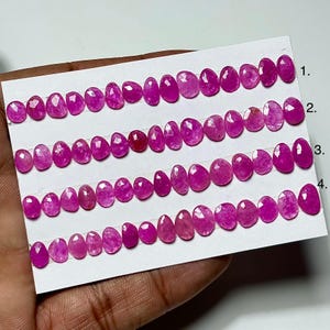 May include: Rows of faceted, oval-shaped, pink gemstones are displayed on a white card. The gemstones vary slightly in size and shade, ranging from light to deep pink. The card is held by a hand, showcasing the gems.