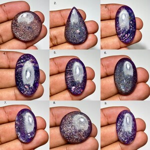 May include: A collection of nine polished gemstones in various shapes, including oval, teardrop, and round. The stones display a mix of purple, silver, and pink hues, with a shimmering, speckled appearance. Each stone is held in a hand, showcasing the natural beauty and unique patterns.
