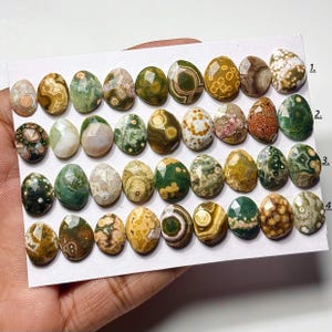May include: A collection of thirty-two faceted, oval-shaped gemstones arranged in four rows on a white card. The stones feature diverse patterns and colors, including shades of green, brown, yellow, and white, with distinct circular and spotted inclusions.