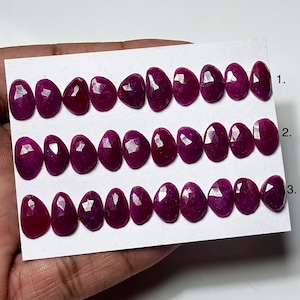 May include: A collection of thirty faceted, teardrop-shaped magenta rubies arranged in three horizontal rows on a white display card. Each row is numbered 1, 2, and 3 on the right side. The gemstones exhibit a polished, reflective surface.