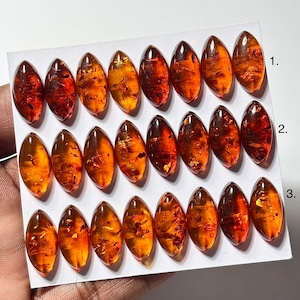 May include: A selection of oval amber gemstones, varying in shades of orange and reddish-brown. The polished stones are arranged in three rows on a white card, highlighting their translucent nature and natural inclusions. Each stone is approximately 2-3 cm in length.