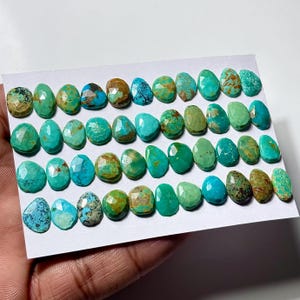 May include: A collection of small, faceted turquoise gemstones arranged in four horizontal rows on a white card. The stones display various shades of blue and green, featuring natural brown matrix patterns and polished, multi-faceted surfaces.