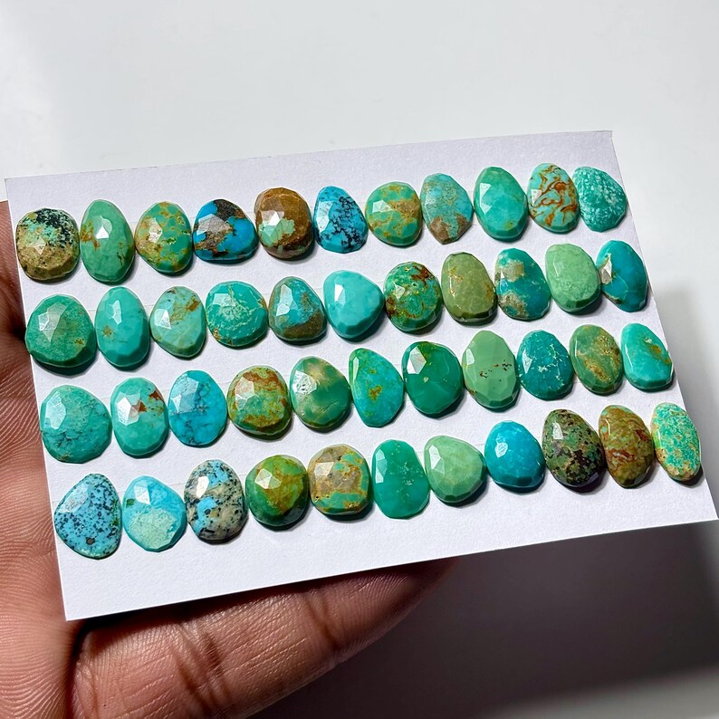 May include: A collection of small, faceted turquoise gemstones arranged in four horizontal rows on a white card. The stones display various shades of blue and green, featuring natural brown matrix patterns and polished, multi-faceted surfaces.