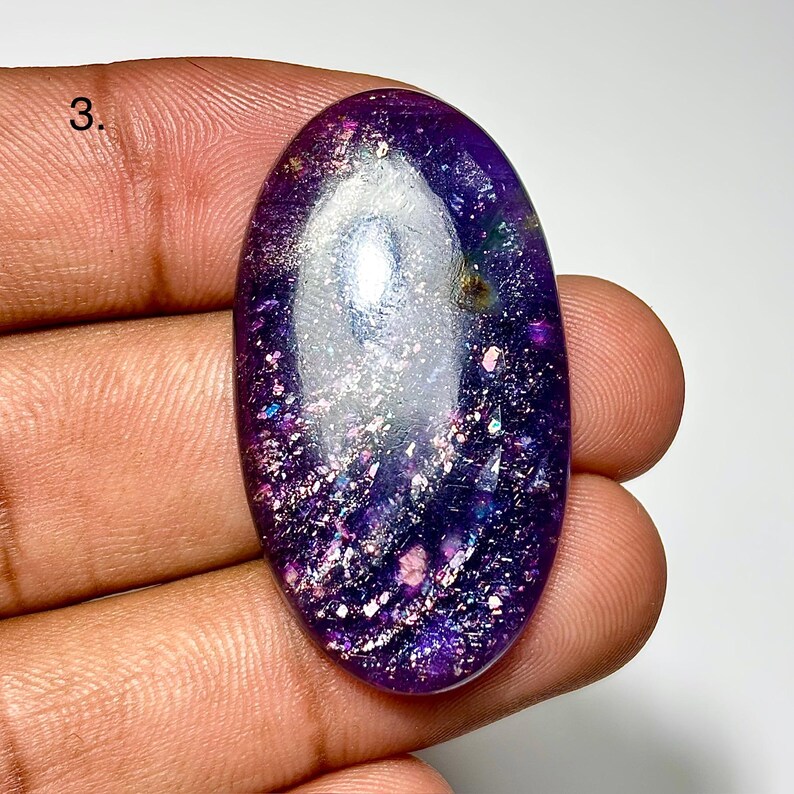 May include: An oval-shaped gemstone with a deep purple hue, speckled with iridescent glitter and flecks of color. The stone is held in a hand, showcasing its polished surface and unique internal patterns. The gemstone's design is eye-catching.