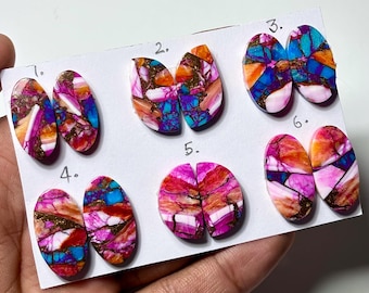 May include: A collection of seven pairs of oval-shaped gemstone cabochons. Each pair features a unique pattern of vibrant colors, including pink, orange, blue, and gold, with a polished finish. The gemstones are displayed on a white card.