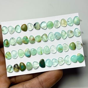 May include: A collection of oval-shaped gemstones in shades of green, teal, and clear, arranged on a white card. The gemstones vary in color and opacity, with some displaying internal patterns. The card is labeled with numbers.
