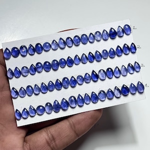 Puede incluir: Filas de gemas azules en forma de lágrima se muestran en una tarjeta blanca. Las gemas tienen un aspecto pulido y translúcido, con variaciones de color y patrones internos. La tarjeta es sostenida por una mano, y las filas están numeradas del 1 al 4.