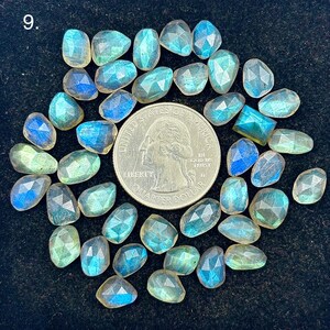 Op de afbeelding: Een verzameling gefacetteerde labradoriet edelstenen in blauwe, groene en gele tinten, gerangschikt rond een Amerikaanse kwart dollar munt. De edelstenen variëren in vorm en grootte, met een gepolijst oppervlak. De achtergrond is donker en getextureerd.