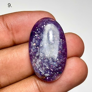 May include: Oval-shaped purple gemstone with glittery inclusions. The polished stone has a translucent quality, with a mix of dark and light purple hues. The gemstone is held in a hand, showcasing its size and shape.