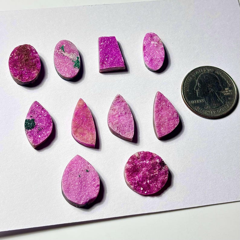 May include: A collection of polished, vibrant pink gemstones in various shapes, including ovals, teardrops, and rectangles. Some stones have green and brown inclusions. A US quarter coin is included for size reference. The gemstones are displayed on a white surface.