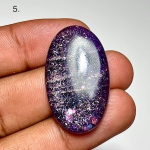May include: An oval-shaped, polished gemstone with a deep purple hue, speckled with iridescent flecks of pink, silver, and blue. The stone is held in a hand, showcasing its smooth, reflective surface. The gemstone is approximately 1.5 inches in length.
