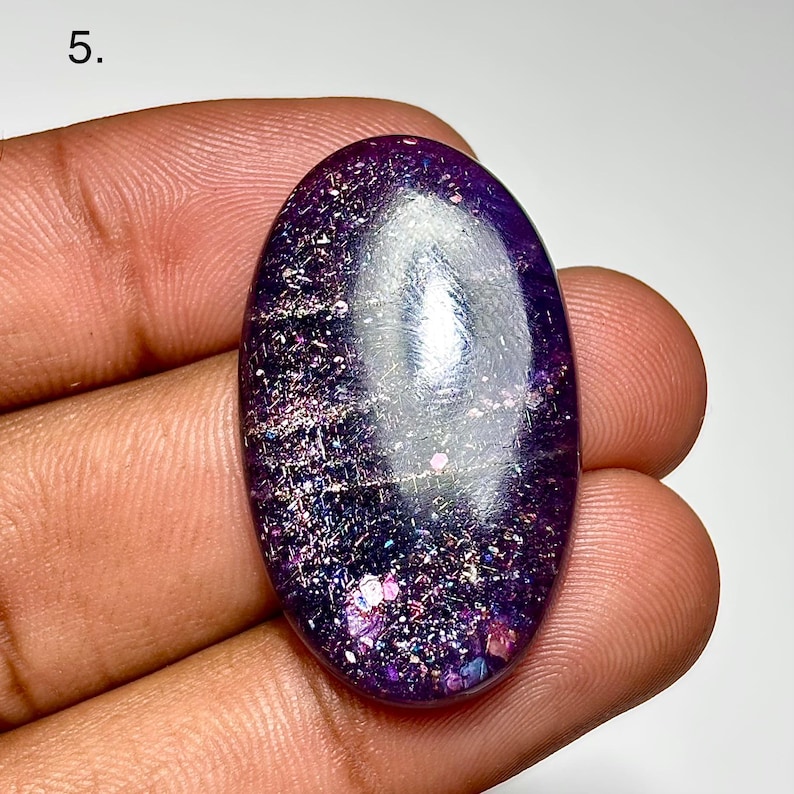 May include: An oval-shaped, polished gemstone with a deep purple hue, speckled with iridescent flecks of pink, silver, and blue. The stone is held in a hand, showcasing its smooth, reflective surface. The gemstone is approximately 1.5 inches in length.