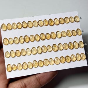 以下が含まれることがあります： 白いカードに、ファセット加工された楕円形の金色の黄色の宝石が並んでいます。宝石はサイズがわずかに異なり、1から4まで番号が付けられています。宝石は研磨された反射面を持っています。