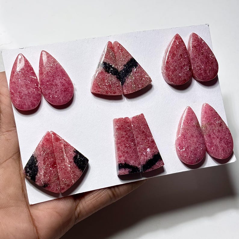 May include: Assortment of pink gemstone cabochons in various shapes, including teardrop and triangular forms. Some stones feature black inclusions, adding contrast to the polished, speckled surfaces. The gemstones are displayed on a white surface.