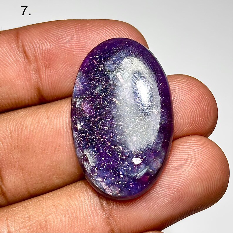 May include: An oval-shaped, polished gemstone with a deep purple hue, speckled with lighter shades and reflective inclusions. The stone is held in a hand, showcasing its smooth, glossy surface. The gem's shape and color suggest it could be used in jewelry.
