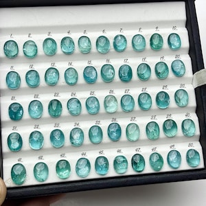 Può includere: Esposizione di cinquanta gemme ovali color teal, disposte in un vassoio nero con scomparti numerati. Le gemme variano leggermente nella tonalità, dal teal chiaro allo scuro. Il vassoio è aperto.