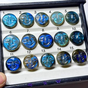 May include: A collection of fifteen round, carved labradorite stones, each featuring a smiling face design. The stones vary in color from blue to green, with visible iridescent flashes. The stones are displayed in a black jewelry box.