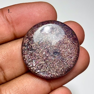 May include: A round, polished gemstone with a shimmering, speckled surface. The gem features a blend of purple, pink, and gold tones, with a clear, reflective area at the top. The overall effect is one of depth and sparkle.