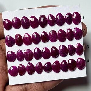 Puede incluir: Una colección de treinta y seis piedras preciosas de rubí en forma de lágrima, facetadas y dispuestas en cuatro filas horizontales sobre una tarjeta blanca. Cada piedra de color magenta intenso presenta una superficie pulida y facetada.