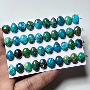 Puede incluir: Colección de piedras preciosas pulidas en varios tonos de azul, verde y marrón. Las piedras, de forma ovalada, están dispuestas sobre una tarjeta blanca. Las piedras tienen un acabado liso y brillante, sujetas en una mano.