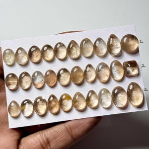 Op de afbeelding: Een verzameling gepolijste, voornamelijk ovaalvormige edelstenen in verschillende tinten amber en lichtbruin. De edelstenen zijn gerangschikt op een witte kaart, met één vierkant geslepen steen. De stenen zijn doorschijnend.