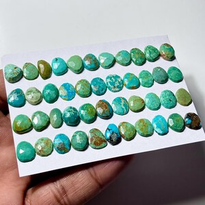 May include: A collection of small, faceted turquoise gemstones arranged in rows on a white card. The stones display various shades of blue and green, featuring natural brown and black matrix patterns, with each piece having a polished, multi-faceted surface.