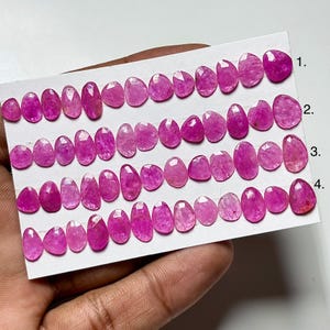 May include: A collection of oval and teardrop-shaped pink gemstones arranged on a white card. The gemstones vary in size and have a polished, faceted surface. The card is labeled with numbers 1 through 4, indicating different rows of stones.