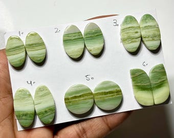 May include: Assortment of six pairs of polished gemstones in various shapes: oval, round, and a unique butterfly-like design. The stones feature a blend of light green and white hues, with visible banding patterns. The gemstones are displayed on a white card.