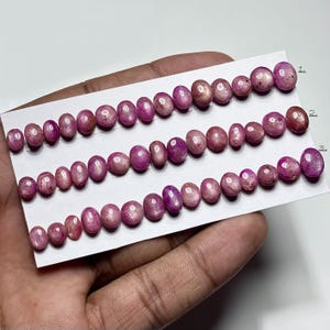 May include: Rows of oval-shaped, polished gemstones in varying shades of pink and purple. The gemstones are arranged on a white card, with each row containing a different number of stones. The stones have a smooth, reflective surface.