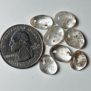 Könnte beinhalten: Sieben kleine, klare, getrommelte Quarzsteine mit sichtbaren inneren Einschlüssen und Rissen, die neben einer amerikanischen 25-Cent-Münze als Größenvergleich angeordnet sind. Die Steine sind glatt, abgerundet und variieren in Form und Größe.