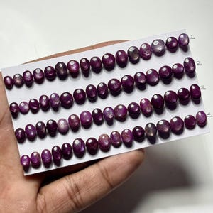 Könnte beinhalten: Eine Sammlung ovaler, tiefroter Edelsteine, die in Reihen auf einer weißen Karte angeordnet sind. Die Edelsteine variieren in der Größe und haben eine polierte, reflektierende Oberfläche. Die Karte wird von einer Hand gehalten, mit den Edelsteinen nummeriert 1 bis 4.