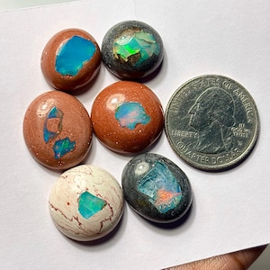 May include: Six oval-shaped gemstones with vibrant colors, including blue, green, and orange, are displayed. The stones are set in a variety of earthy tones, such as brown, gray, and white. A quarter coin is next to the stones for size comparison.