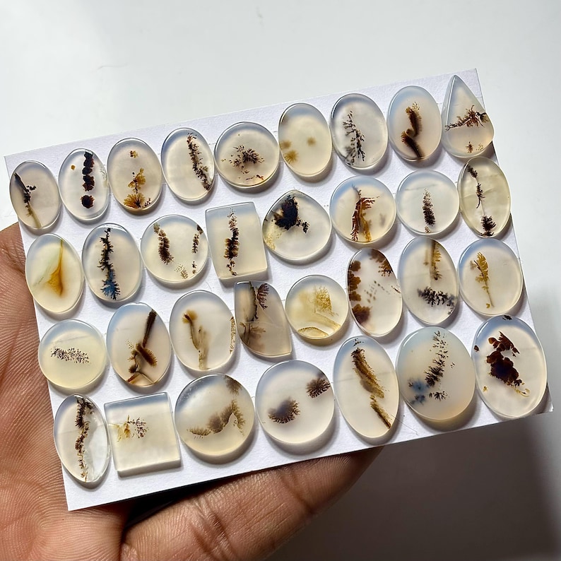Puede incluir: Una colecci&oacute;n de piedras preciosas de &aacute;gata pulidas en varias formas, incluyendo ovaladas, rectangulares y en forma de l&aacute;grima. Las piedras transl&uacute;cidas presentan intrincados patrones de inclusiones marrones, negras y amarillas, sobre un fondo blanco lechoso. Las piedras preciosas se muestran sobre una superficie blanca.