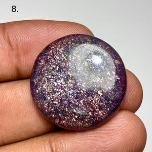 May include: A round, polished gemstone with a deep purple base color and shimmering, glitter-like inclusions. The surface reflects light, creating a sparkling effect. The gemstone is held in a hand, showcasing its size and shape.