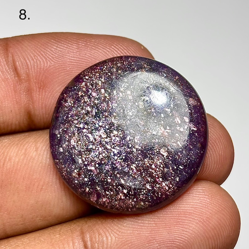 May include: A round, polished gemstone with a deep purple base color and shimmering, glitter-like inclusions. The surface reflects light, creating a sparkling effect. The gemstone is held in a hand, showcasing its size and shape.