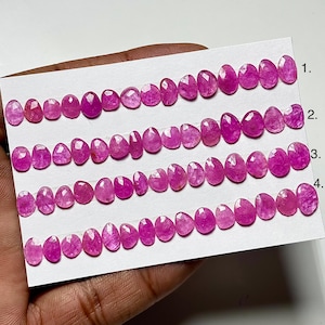 May include: Four strands of oval-shaped, vibrant pink gemstones are displayed on a white card. Each strand is numbered, showcasing the gemstones' variations in size and cut. The gems have a polished, translucent appearance.