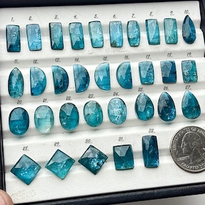 May include: A selection of teal gemstones in diverse shapes, such as rectangles, ovals, and teardrops, presented in a black tray. A UK coin is included for size reference. The gemstones are faceted, reflecting light.