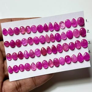 May include: A collection of small, vibrant pink gemstones arranged in four horizontal rows on a white card. The stones feature various shapes, including oval, round, and teardrop cuts, all displaying a consistent deep magenta hue and polished finish.