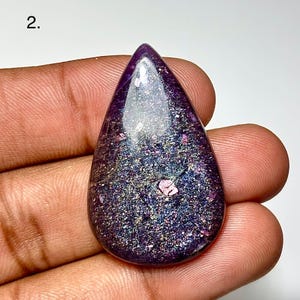 May include: A teardrop-shaped, polished gemstone with a deep purple hue. The stone has a shimmering, glittery appearance with flecks of iridescent color. The top of the stone is a darker shade of purple, and the bottom is a lighter, more sparkly color.
