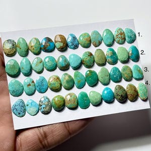 May include: A collection of loose, faceted turquoise gemstones arranged in four horizontal rows on a white card. The stones vary in shades of blue and green, featuring unique brown and black matrix patterns. The rows are numbered 1 through 4.