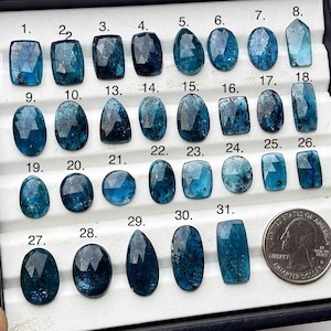 May include: A collection of 31 faceted, teal-colored gemstones in various shapes, including oval, teardrop, and rectangular cuts. A US quarter is included for size reference. The gemstones are displayed in a black tray.