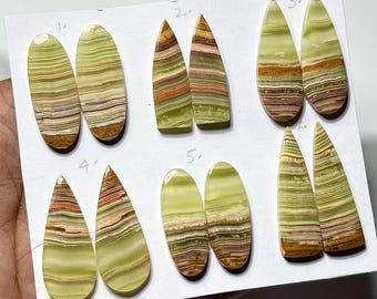 May include: A collection of six pairs of polished onyx gemstones in various shapes. The stones display a layered pattern of green, brown, and reddish-brown hues. Each pair is numbered for identification, showcasing the natural variations in color and design.