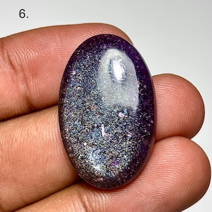 May include: An oval-shaped, polished gemstone with a deep purple hue. The stone has a shimmering, speckled appearance with iridescent flecks of blue, green, and silver. The number "6." is visible in the upper left corner.