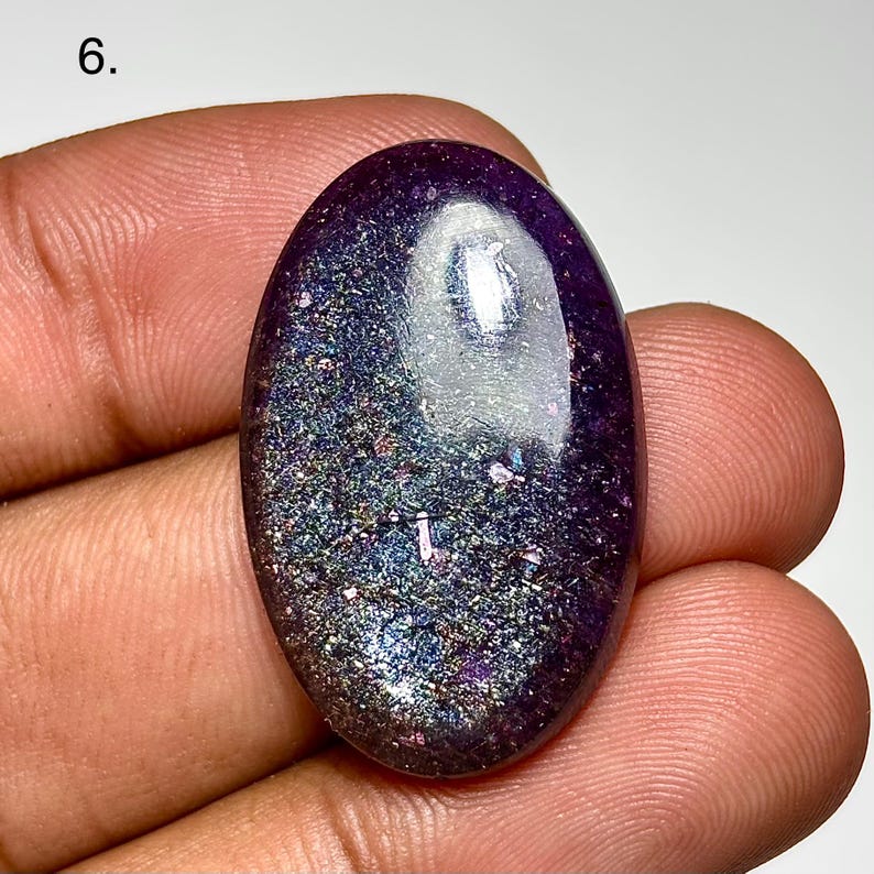 May include: An oval-shaped, polished gemstone with a deep purple hue. The stone has a shimmering, speckled appearance with iridescent flecks of blue, green, and silver. The number "6." is visible in the upper left corner.