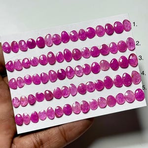 Könnte beinhalten: Reihenweise ovale, facettierte, rosa Edelsteine auf einer weißen Karte. Die Steine, vermutlich Rubine, variieren in der Farbe von hell- bis tiefrosa. Geeignet für die Schmuckherstellung.